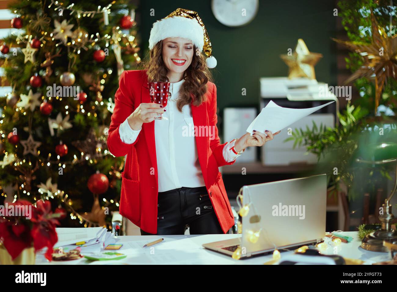 Noël. Heureuse femme élégante propriétaire de petite entreprise dans le chapeau de santa et veste rouge avec des documents et du verre de vin dans le bureau vert moderne avec Christma Banque D'Images