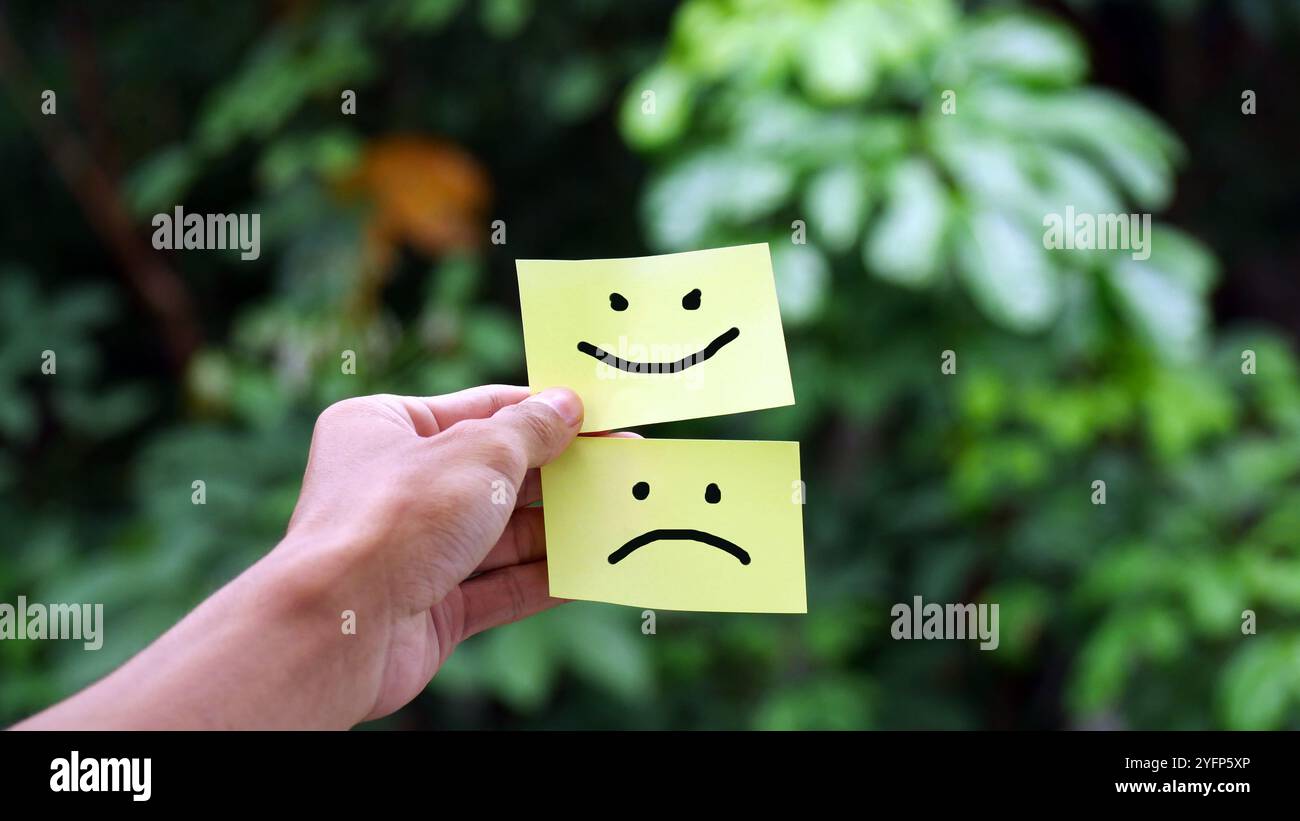 Mains tenant des cartes de visage tristes et souriantes sur fond de nature floue. Concept de sautes d'humeur, personnalité divisée et santé mentale Banque D'Images