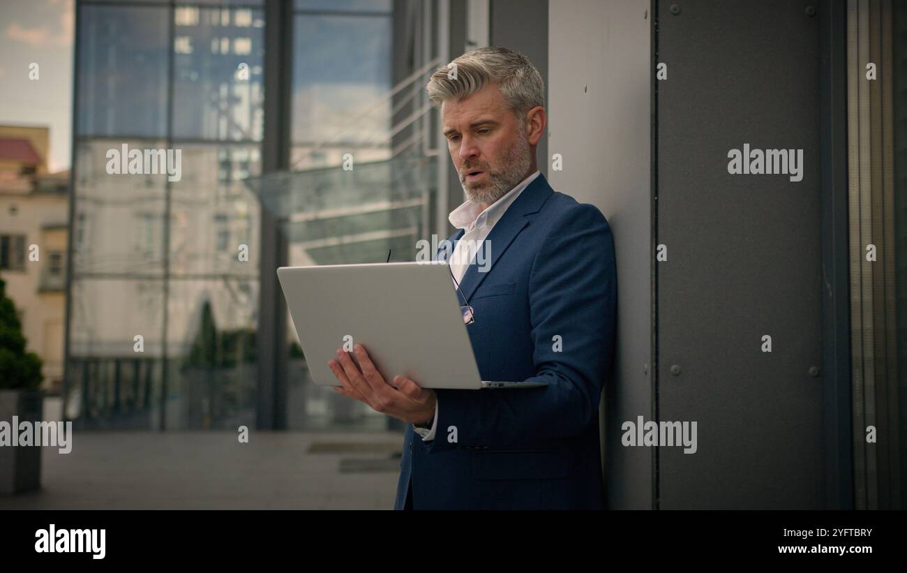 Caucasien âgé homme senior gestionnaire d'affaires mature occupé homme d'affaires d'employeur d'âge moyen penchant bâtiment de bureau de mur tapant ordinateur de travail portable Banque D'Images