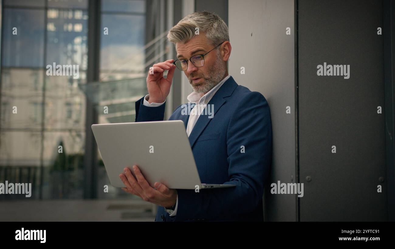 Caucasien âgé homme senior gestionnaire d'affaires mature occupé homme d'affaires d'employeur d'âge moyen penchant bâtiment de bureau de mur tapant ordinateur de travail portable Banque D'Images