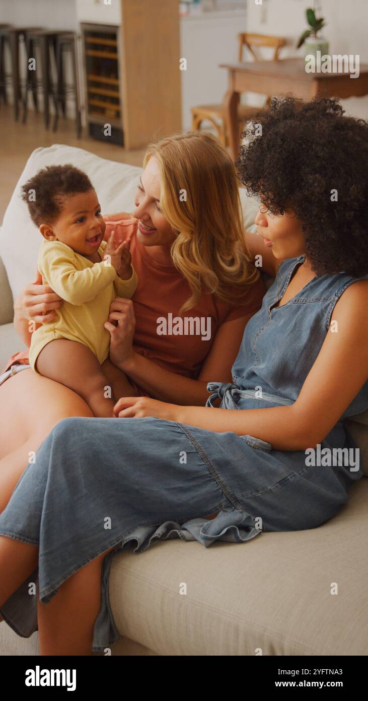Deux femmes jouent joyeusement avec un adorable bébé assis sur un canapé confortable dans un salon lumineux et accueillant. Leurs expressions radiat Banque D'Images