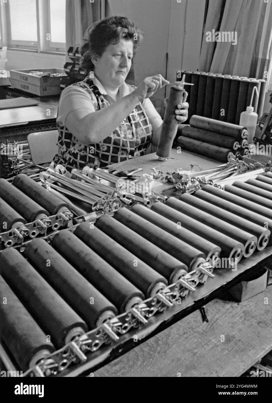 Une femme insérant des manchons extérieurs en caoutchouc sur des cylindres métalliques, dans une usine d'ingénierie Vickers vers 1970. Vickers était une société d'ingénierie britannique qui a existé de 1828 à 1999. Elle a commencé à Sheffield comme fonderie d'acier, célèbre pour le moulage des cloches d'église. Elle s'est diversifiée dans l'équipement militaire et la construction navale. En 1911, elle s'est développée dans la fabrication d'avions, puis dans l'électricité et les chemins de fer. Après la seconde Guerre mondiale, Vickers-Armstrong fabriqua des avions commerciaux, dont le VC10 en 1959. Dans les années 1960, des parties de Vickers ont été nationalisées, et en 1999, le reste a été acquis par Rolls-Royce. Banque D'Images