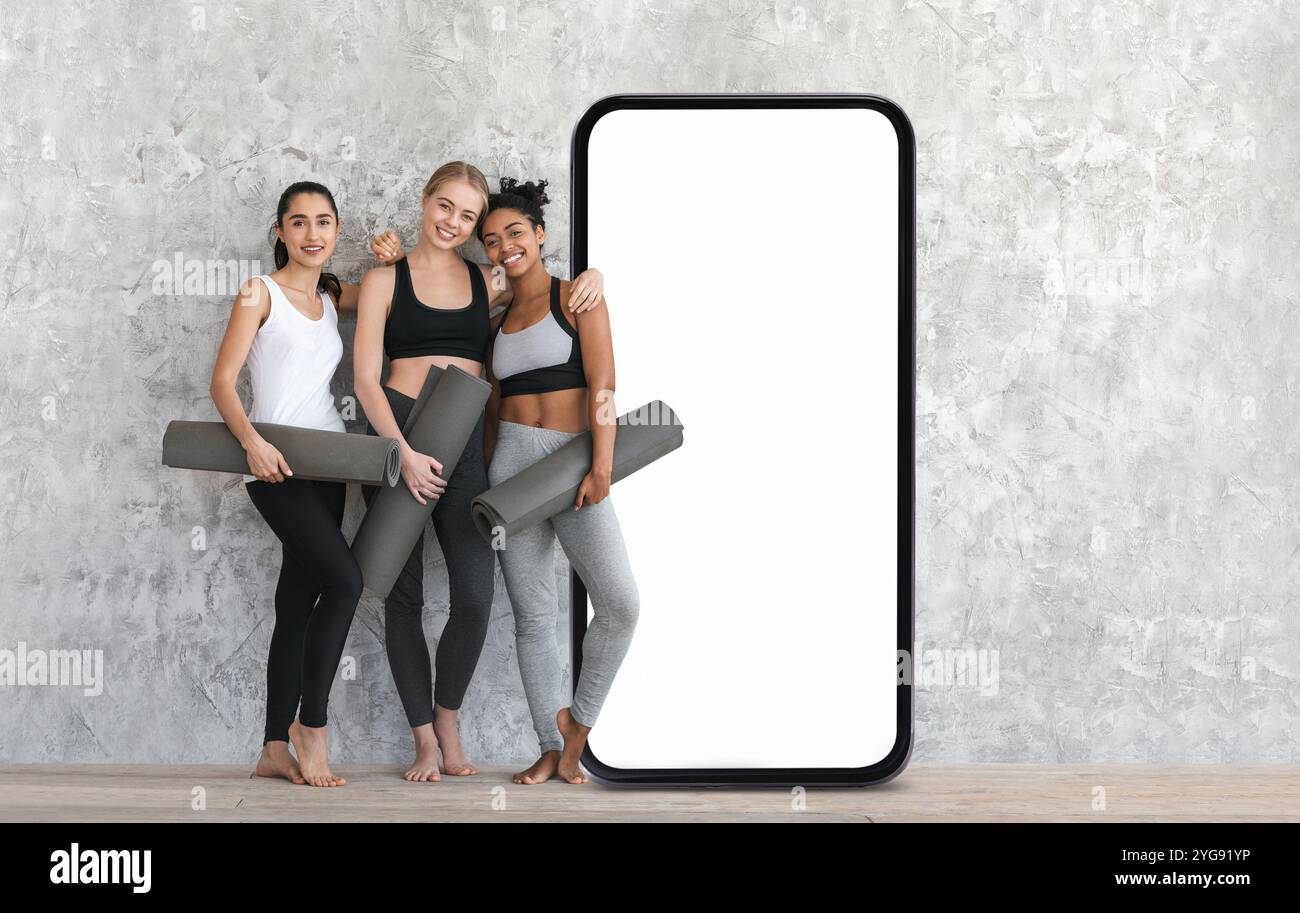Trois femmes tenant des tapis de yoga près d'un écran de téléphone vierge dans un studio Banque D'Images