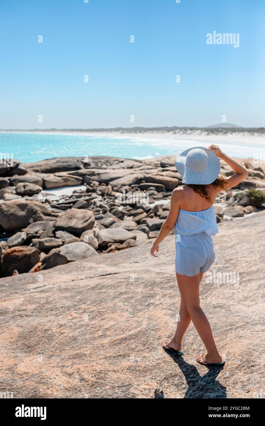 Jeune femme brune sur des vacances d'été tenant un chapeau blanc à motifs, marchant à mi-foulée sur les rochers. Banque D'Images