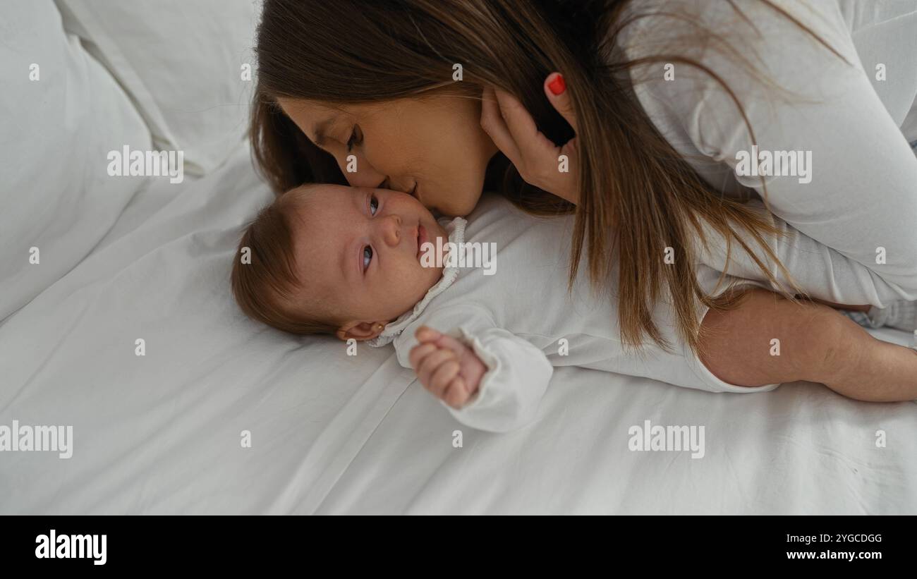 Mère embrasse amoureusement sa petite fille tout en étant couchée ensemble sur un lit blanc dans une chambre confortable, capturant un moment familial tendre rempli d'affection Banque D'Images