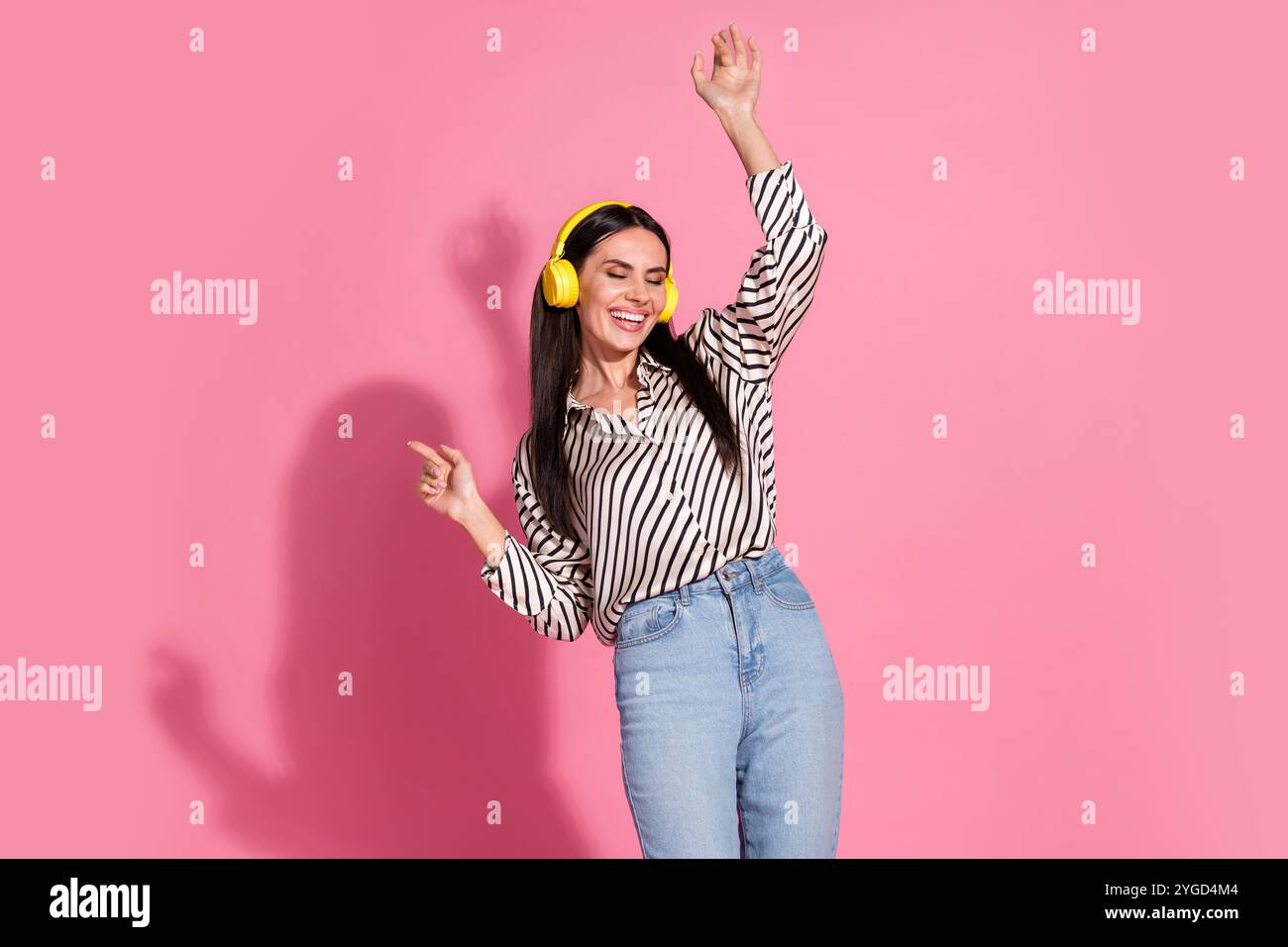 Jeune femme en chemise rayée appréciant la musique avec des écouteurs jaunes sur fond rose Banque D'Images