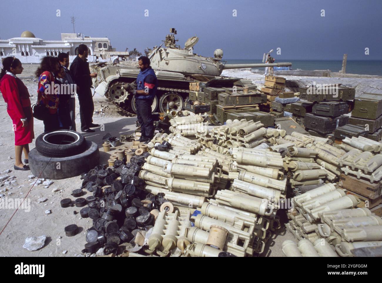 Première Guerre du Golfe : 10 mars 1991 Un soldat koweïtien se tient parmi un énorme stock de bombes de mortier irakiennes abandonnées et de mines antipersonnel sur la plage d'Anjafa dans la ville de Koweït. Il montre une bombe de mortier à un groupe de curieux Koweïtiens locaux. À côté d'eux se trouve un char chinois type 69 dans son revêtement. Banque D'Images