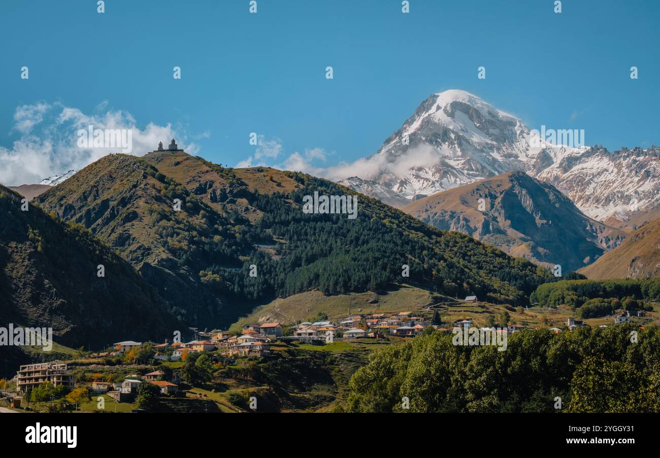 Gergeti Trinity Church perchée sur une colline avec le mont Kazbek enneigé dominant en arrière-plan, et le village de Stepantsminda en bas Banque D'Images