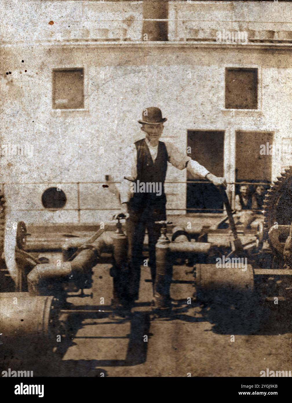 Cette photographie vintage représente un homme en tenue du XIXe siècle, y compris un chapeau melon, debout à côté de grands engrenages et tuyaux sur ce qui semble être le pont d'un bateau à vapeur. Le cadre maritime et ses vêtements suggèrent une période de la fin des années 1800 au début des années 1900, probablement pendant le pic des navires à vapeur. L'image offre un aperçu rare de la technologie maritime historique et de la vie sur les navires, préservant un morceau de l'histoire industrielle et maritime de l'époque. Banque D'Images