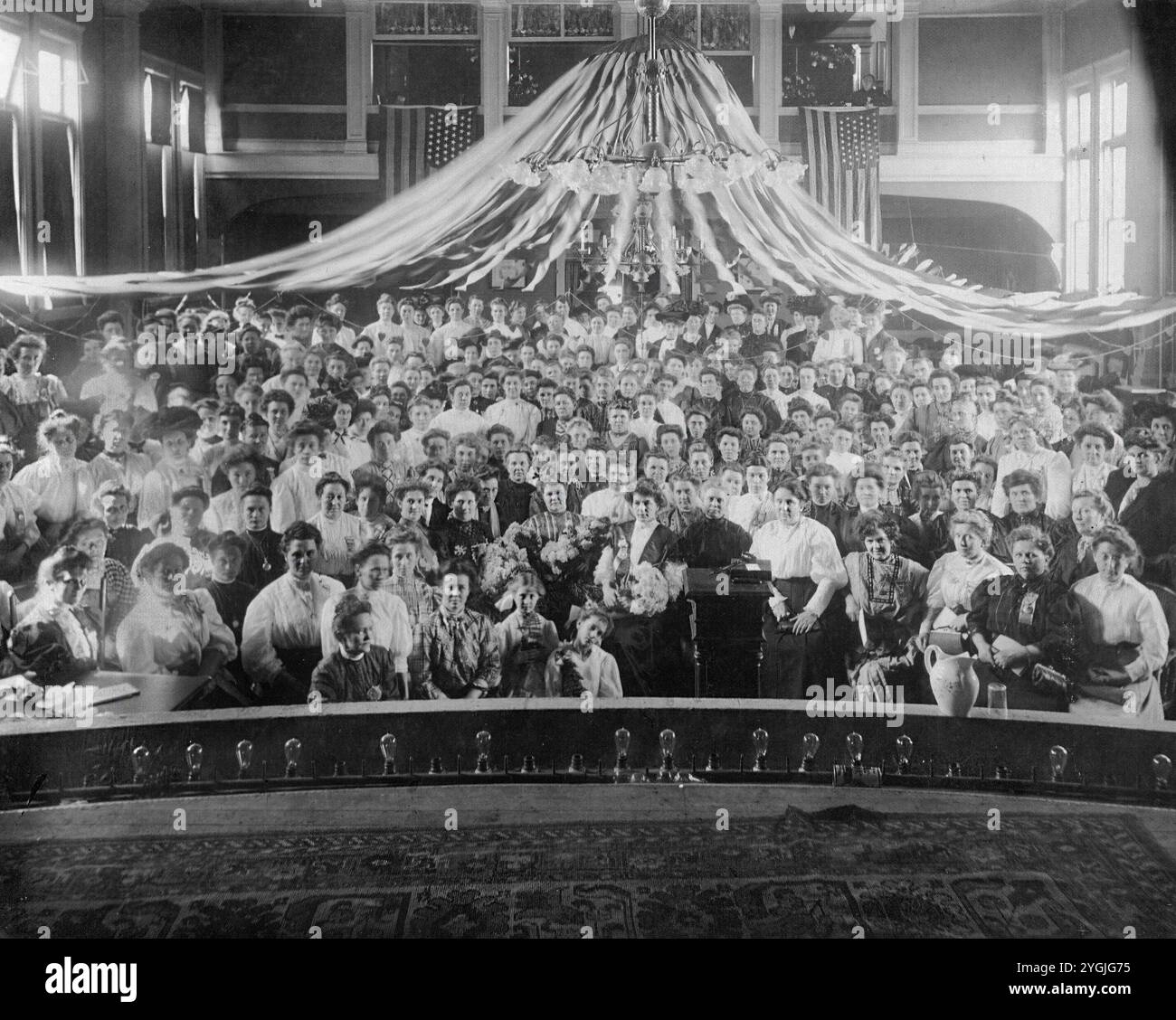 Ceci est une photo originale et réelle d'un grand rassemblement de femmes au début du XXe siècle en Amérique, probablement de l'époque édouardienne (vers 1900-1915). La salle est décorée de drapeaux et de banderoles américains, suggérant un événement patriotique ou formel. La tenue vestimentaire des femmes reflète le style de l'époque, capturant un moment d'engagement communautaire et social dans la société américaine. Banque D'Images