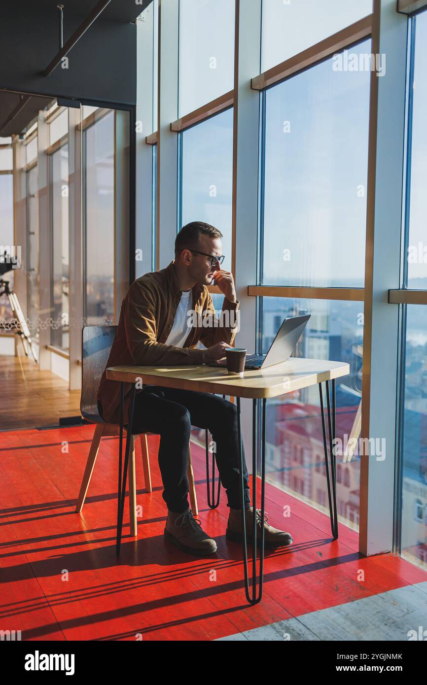 Un homme équilibré dans des lunettes et des vêtements décontractés travaille sur un ordinateur portable à partir d'un espace de travail confortable. Un freelance performant travaille à distance. Banque D'Images