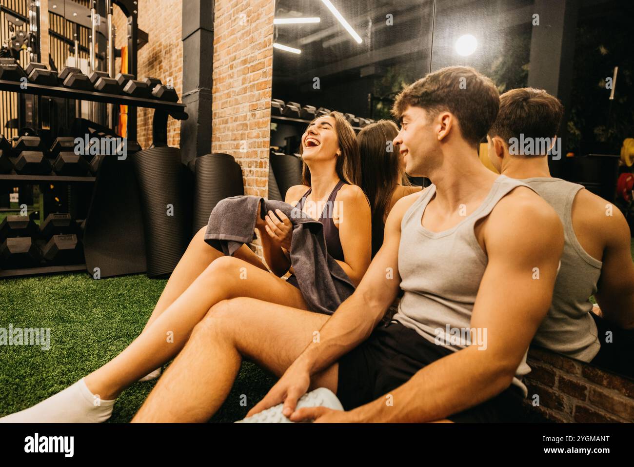 Un jeune couple profite d'un moment de légèreté tout en étant assis sur le sol de la salle de sport. Ils rient et semblent détendus après une séance d'entraînement. Banque D'Images