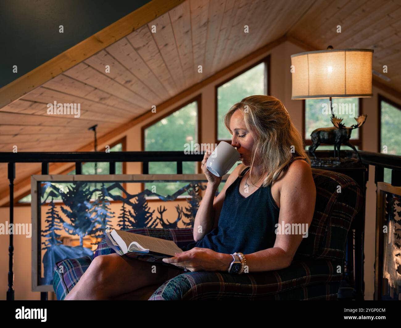 Une femme est assise confortablement dans une cabine confortable, sirotant une boisson chaude tout en lisant son livre par les fenêtres. Banque D'Images