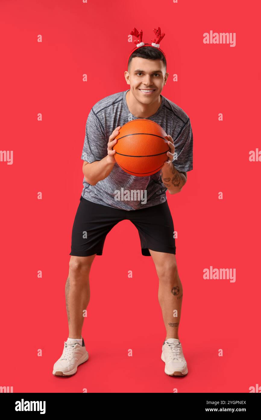 Jeune homme heureux dans des cornes de renne jouant au basket-ball sur fond rouge Banque D'Images