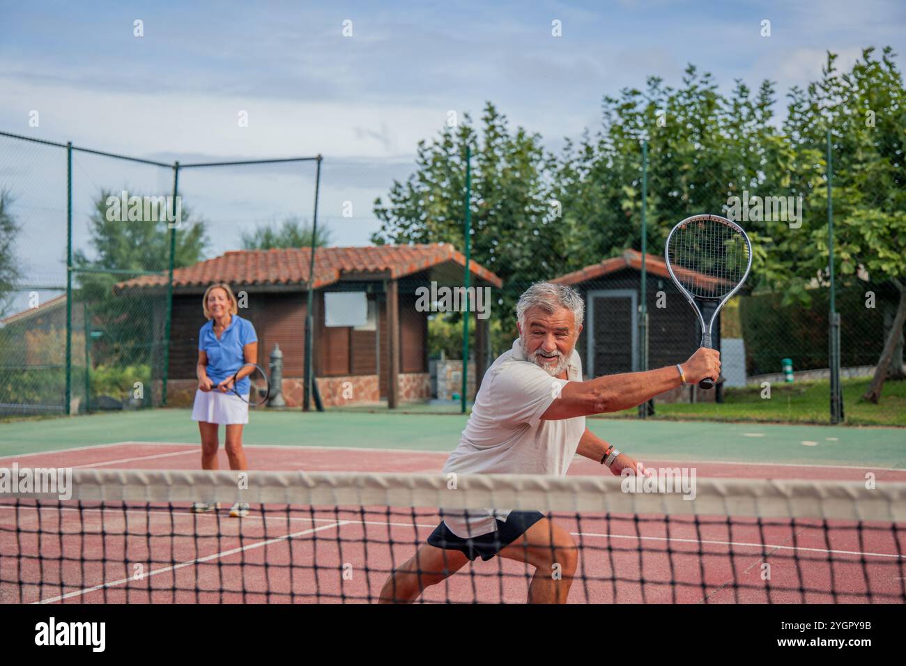 un joueur de tennis senior exécute un tir du revers pendant un match, avec son partenaire debout à côté de lui sur le court de tennis. Leurs mouvements synchronisés h Banque D'Images