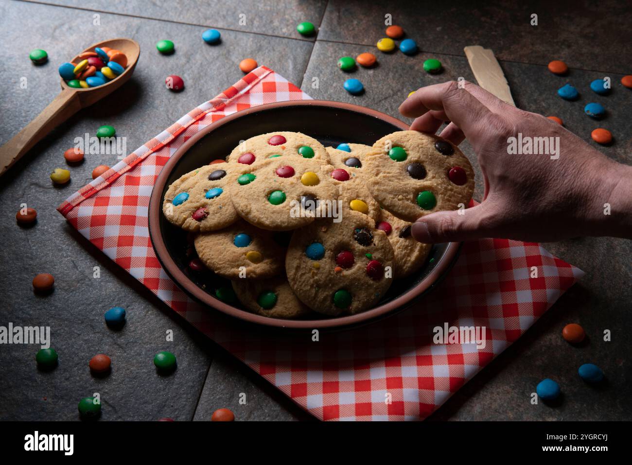 Main de l'homme prenant un biscuit à la vanille avec des pépites de chocolat rouge, bleu, vert et jaune d'un groupe de biscuits sur une plaque noire ronde sur une serviette rouge Wit Banque D'Images