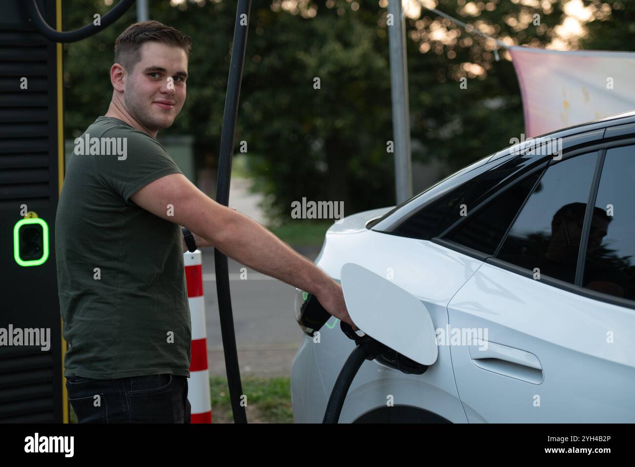 Un homme sourit tout en chargeant sa voiture électrique à une station de recharge publique, montrant un style de vie positif et respectueux de l'environnement. Banque D'Images