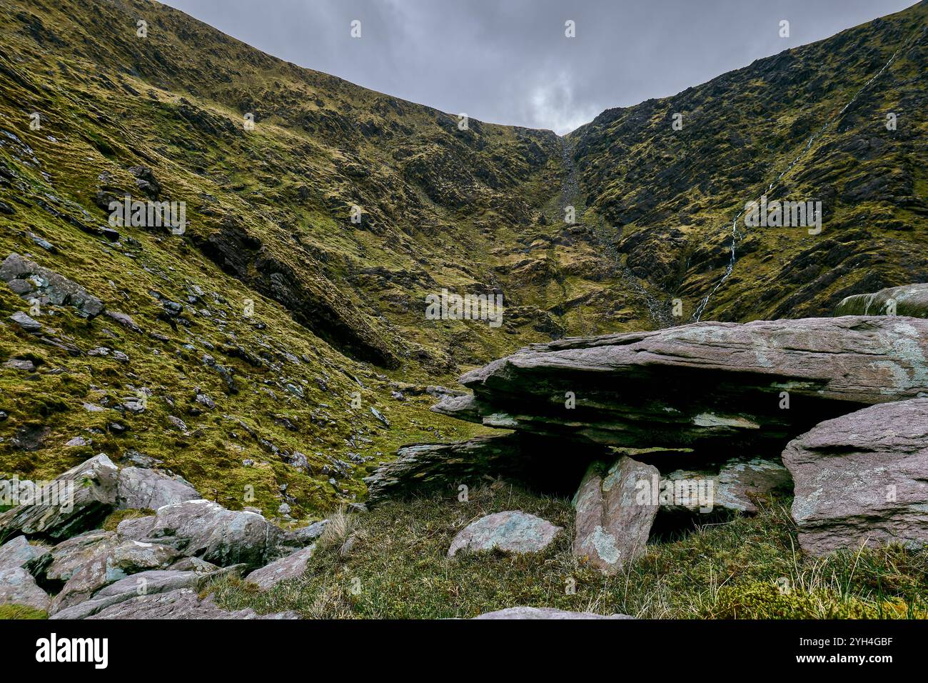 Une vallée tranquille présente de grands rochers couverts de mousse entourés de falaises abruptes sous un ciel nuageux, soulignant la végétation luxuriante de la colline isolée Banque D'Images