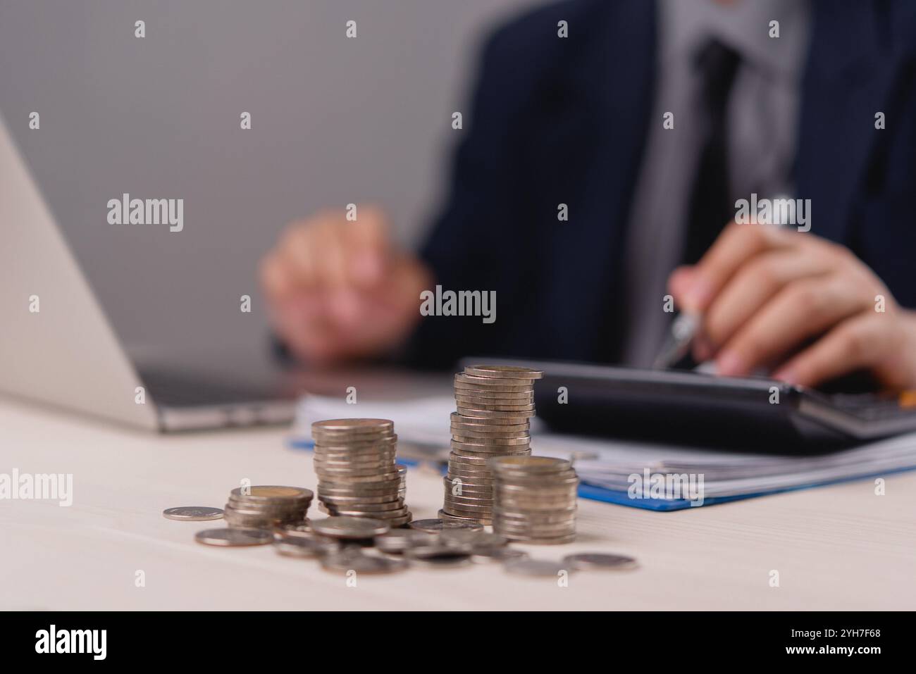 Pièces empilées sur un bureau au premier plan avec un homme d'affaires calculant les finances en arrière-plan, symbolisant l'épargne, l'investissement, et ma financière Banque D'Images