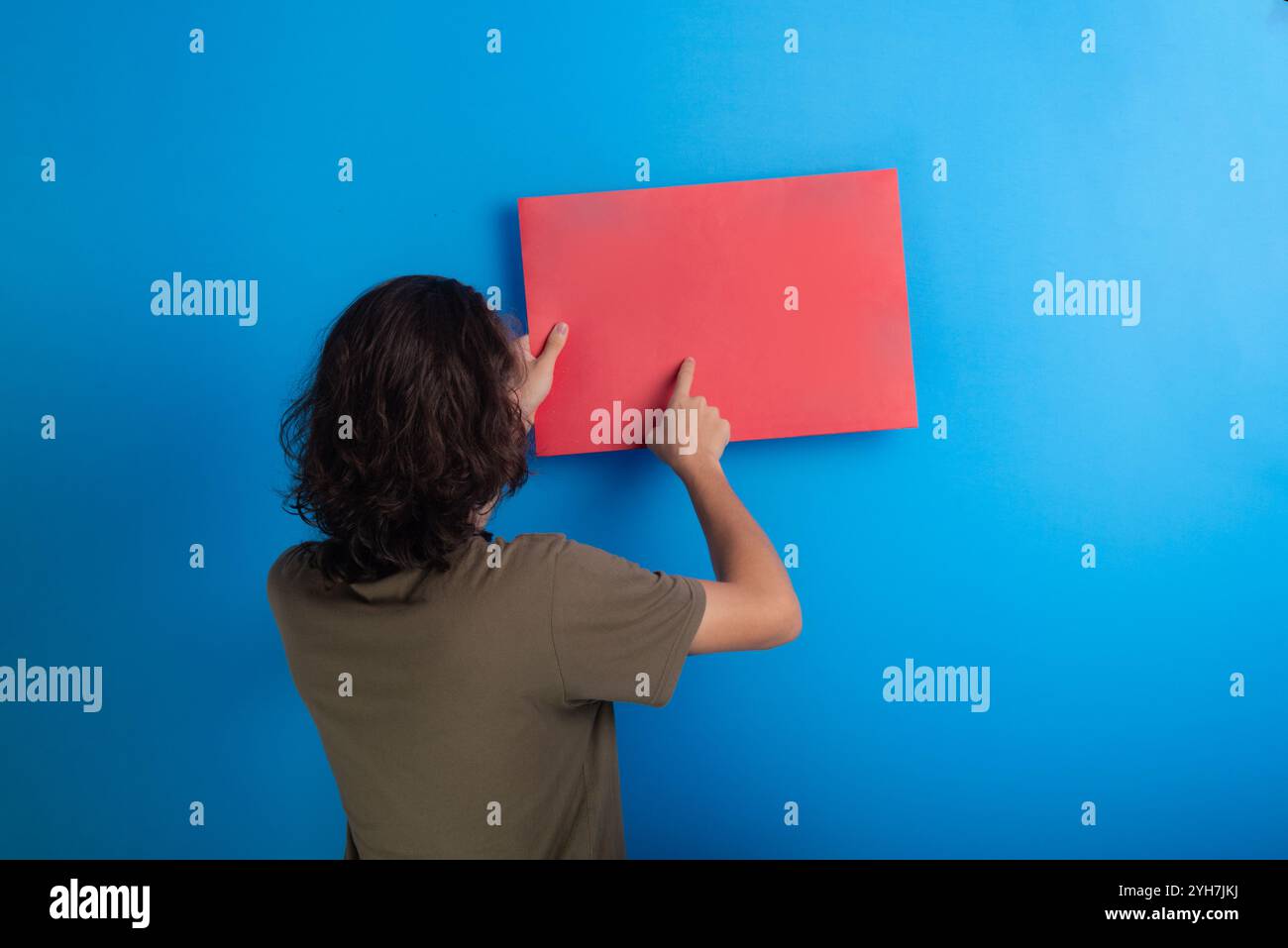 jeune homme tenant une affiche vide de couleur rouge. Confiant et pointant le doigt vers le centre de l'affiche. Isolé sur fond bleu. Banque D'Images