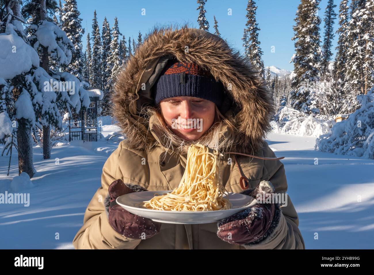 Femme debout à l'extérieur dans le paysage arctique froid du nord pendant le jour de température glaciale -40 tenant une assiette de nouilles de pâtes congelées avec fourchette. Banque D'Images