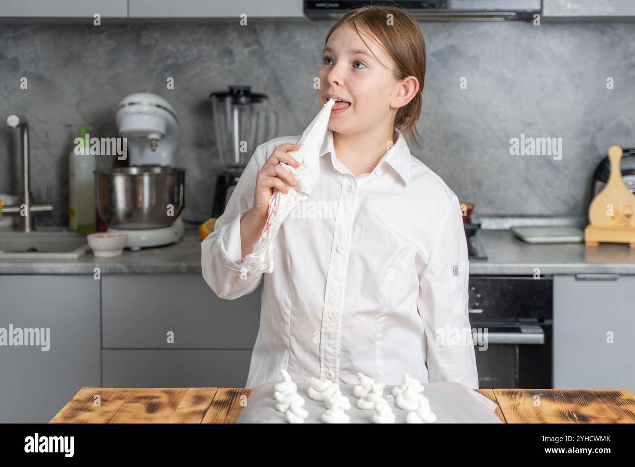 Une petite fille mignonne dans la cuisine prépare la meringue dans une robe de chef, elle tient un sac à pâtisserie et lèche la meringue restante. Photo de haute qualité Banque D'Images