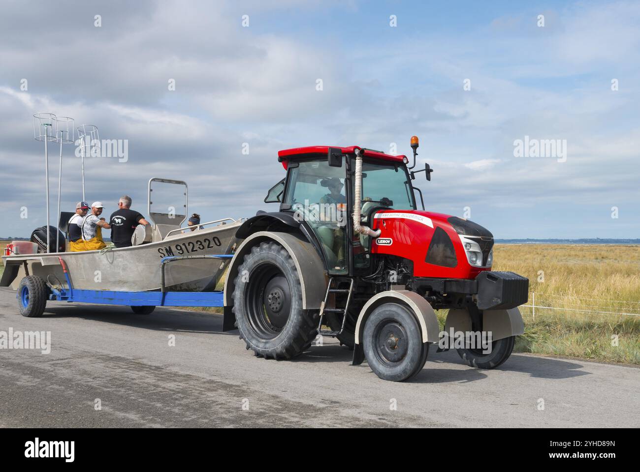 Tracteur rouge avec remorque et gens sur une route de campagne, entouré de champs, tracteur avec bateau sur le chemin de la récolte des moules, Cherrueix, Bretagne, Banque D'Images