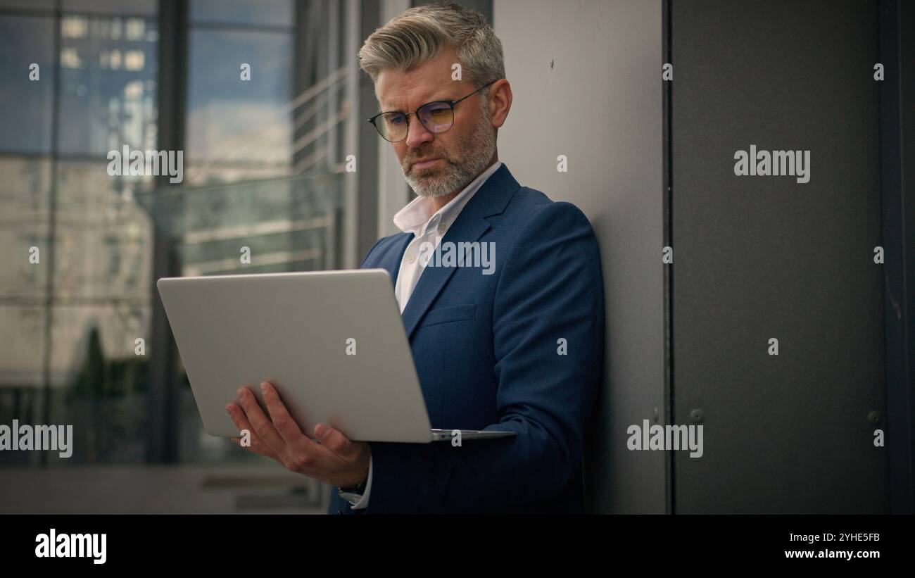 Caucasien âgé homme senior gestionnaire d'affaires mature occupé homme d'affaires d'employeur d'âge moyen penchant bâtiment de bureau de mur tapant ordinateur de travail portable Banque D'Images