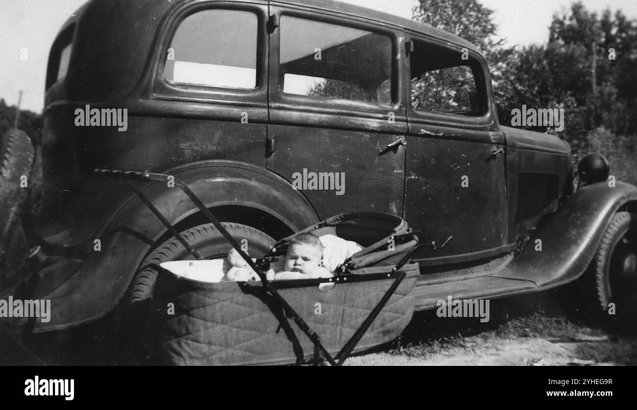 Un bébé chubby est assis dans un berceau à côté de la voiture familiale, vers 1930. Banque D'Images