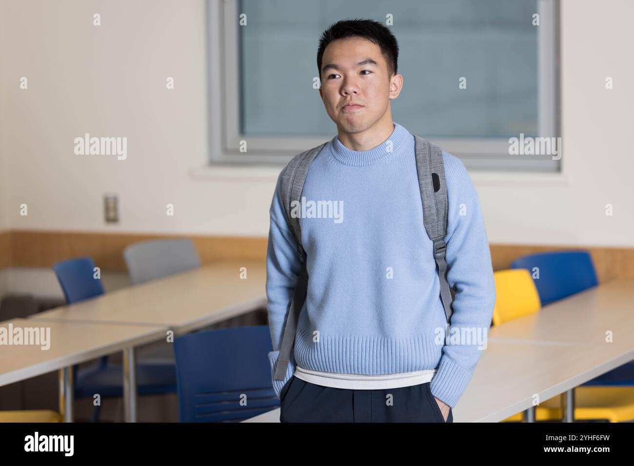 Un jeune étudiant universitaire asiatique debout dans une salle de classe avec son sac à dos devant des bureaux et des chaises Banque D'Images