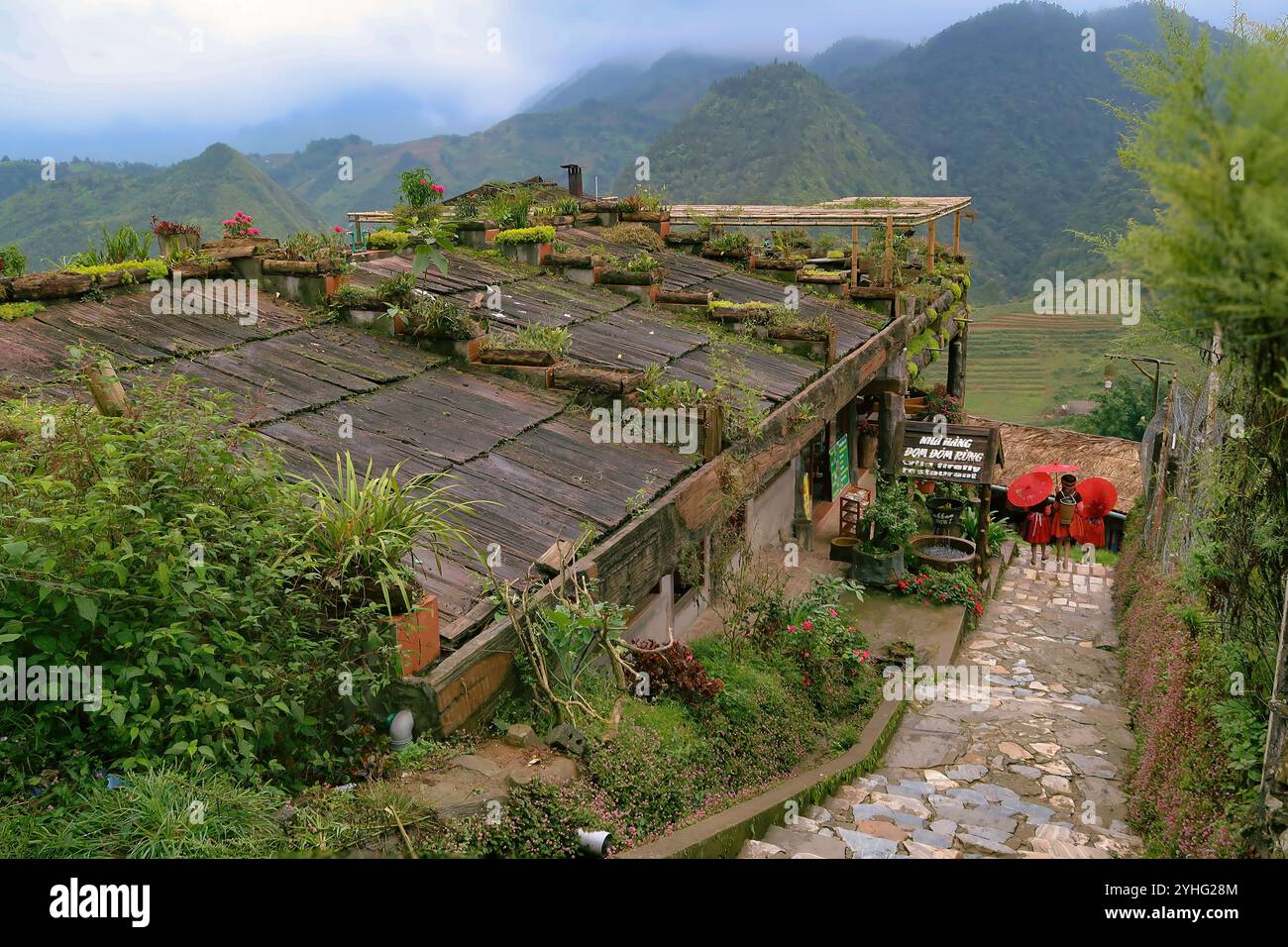 Une maison vietnamienne traditionnelle avec un toit vert, entourée de montagnes et de fleurs colorées, avec deux femmes tenant des parapluies. Banque D'Images