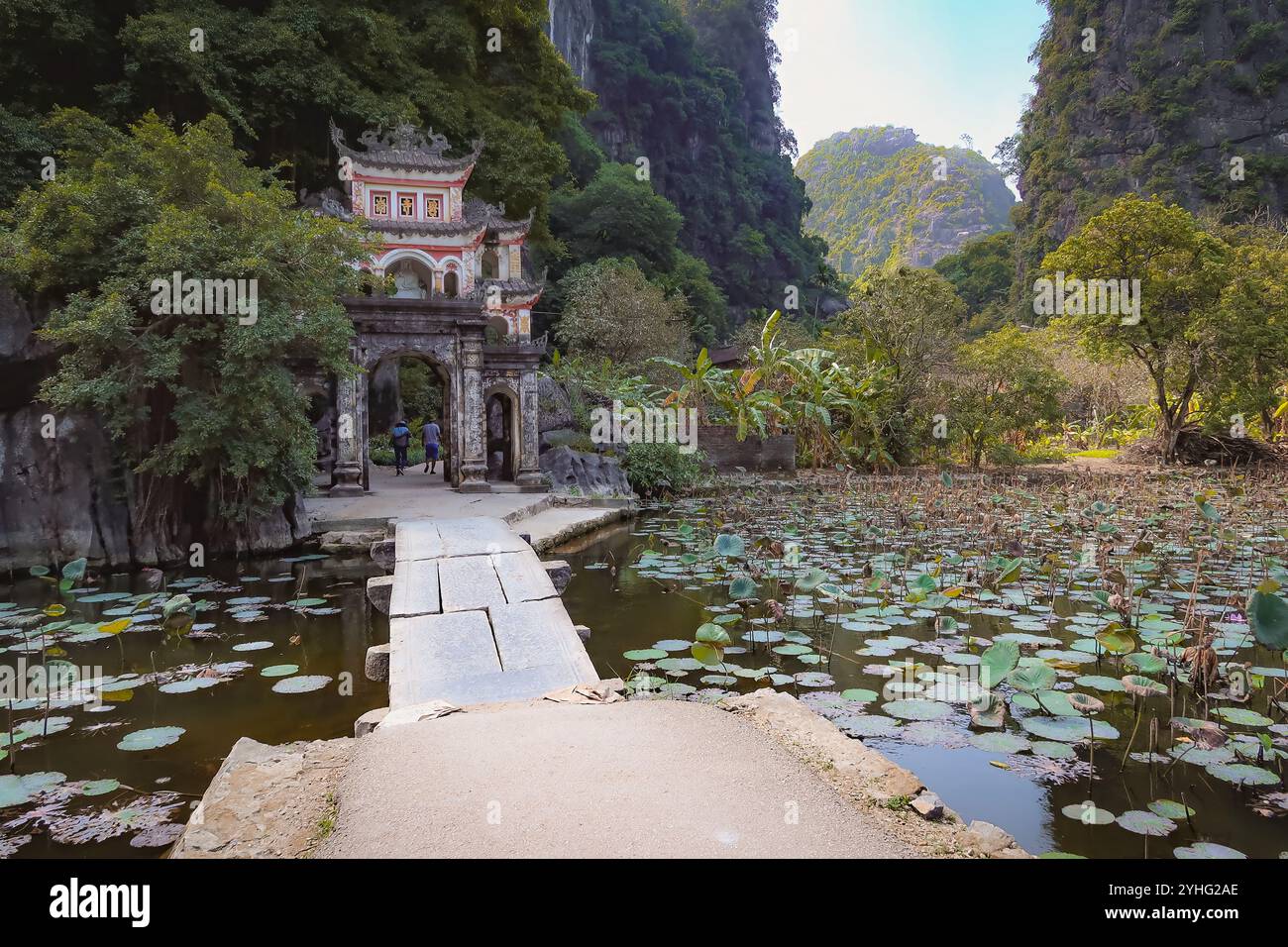 Une vue sereine sur une porte vietnamienne traditionnelle à la pagode Bich Dong, avec un chemin de pierre menant à un étang de lotus, entouré de falaises verdoyantes. Banque D'Images