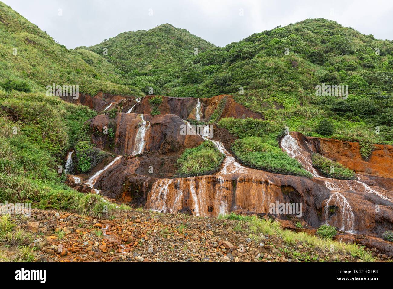 Une petite cascade en bord de route à Juifen, Taiwan, avec de l'herbe verte luxuriante et des roches orange / brunes contrastées, l'eau en cascade dans des rivets blancs. Banque D'Images