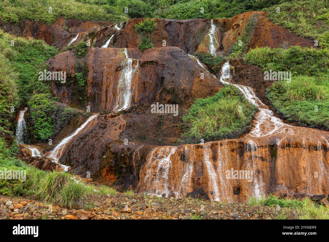 Une petite cascade en bord de route à Juifen, Taiwan, avec de l'herbe verte luxuriante et des roches orange / brunes contrastées, l'eau en cascade dans des rivets blancs. Banque D'Images