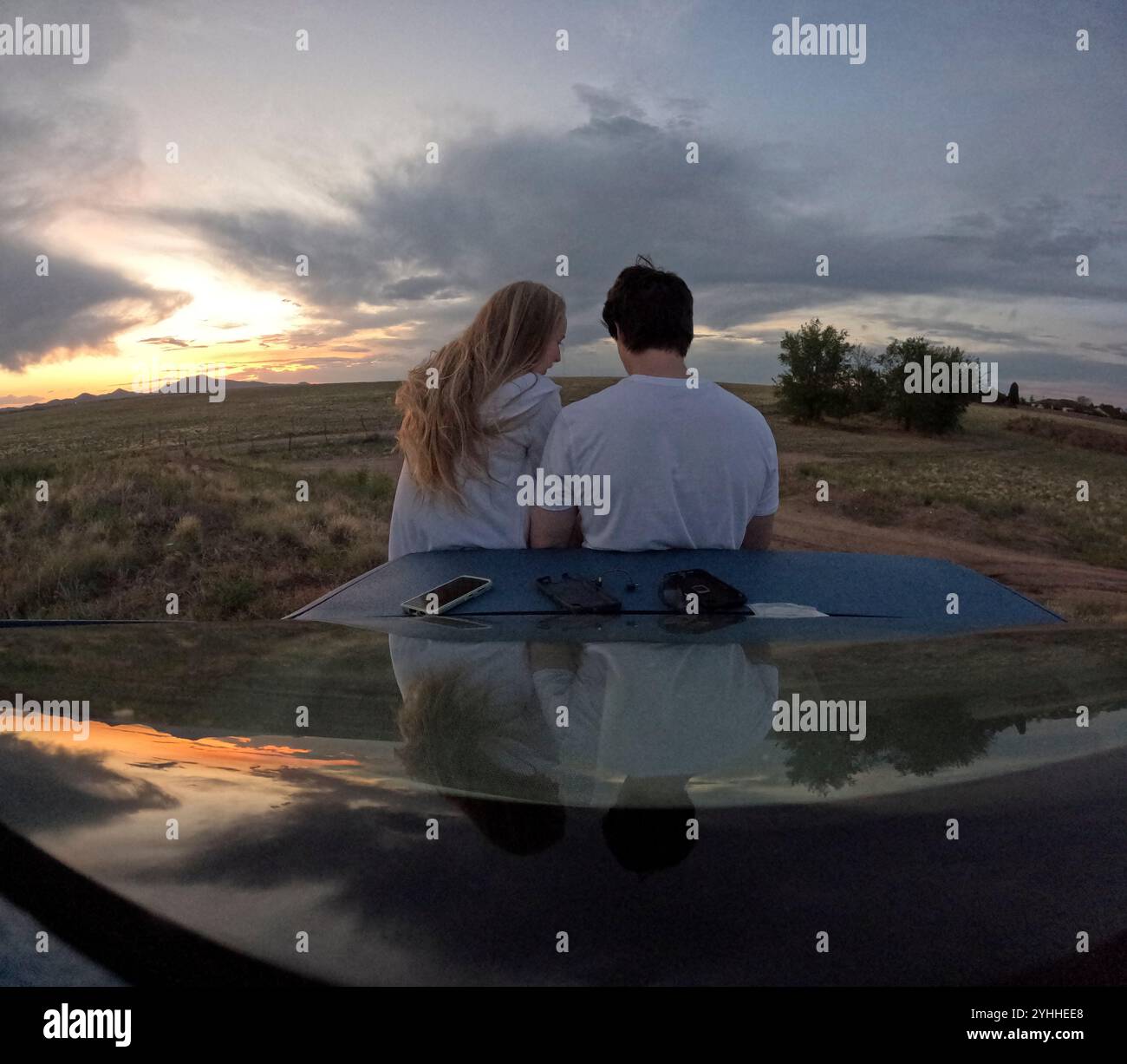 Couple profite d'un coucher de soleil serein tout en étant assis sur un capot de voiture dans un paysage rural Banque D'Images