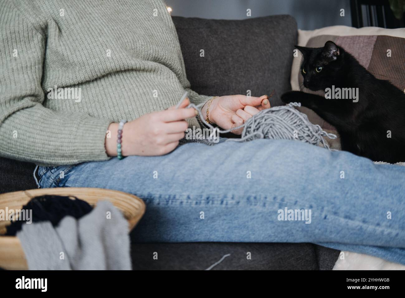 Femme tricotant avec chat noir sur canapé, embrassant temps d'arrêt confortable. Relaxation, compagnie, détox numérique et concept de passe-temps attentif. Banque D'Images