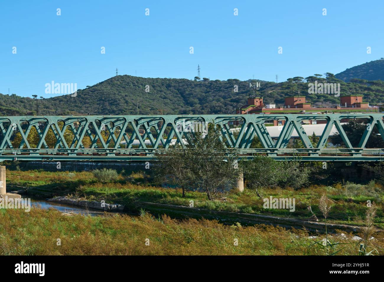 Montcada i Reixac. Espagne - 12 novembre 2024 : un pont historique en treillis d'acier vert enjambe la rivière Ripoll, avec des collines verdoyantes et un ciel bleu clair pr Banque D'Images