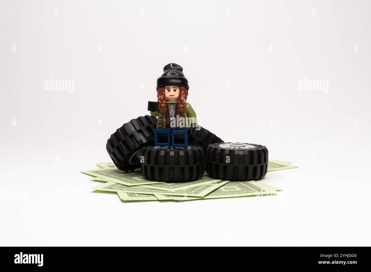 Fille Lego miniature dans la tenue d'hiver assis sur des pneus de voiture empilés sur des billets d'un dollar. Banque D'Images