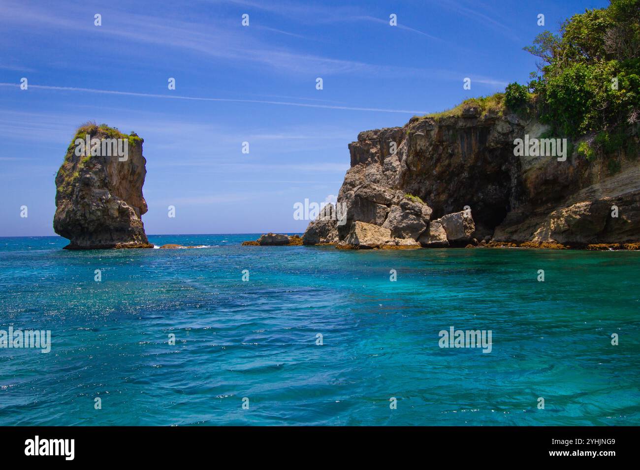 De douces vagues se glissent contre des falaises accidentées et une imposante formation rocheuse sous un ciel bleu éclatant. Une végétation luxuriante domine le paysage côtier, ajoutant un touc Banque D'Images