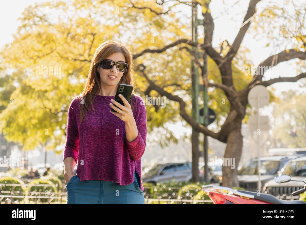 Élégante femme d'âge moyen sourit alors qu'elle regarde son smartphone dans un parc au coucher du soleil. Portant des lunettes de soleil et un pull violet, elle garde une main en lui Banque D'Images