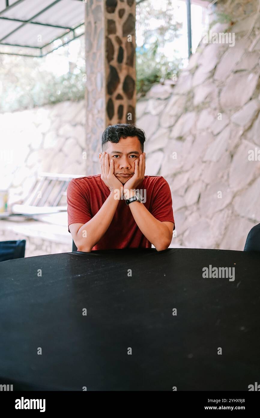 Un homme portant un t-shirt rouge est assis réfléchi à une table noire, reposant son menton sur sa main. Le cadre extérieur avec des murs en pierre et un éclairage naturel ajoutent Banque D'Images