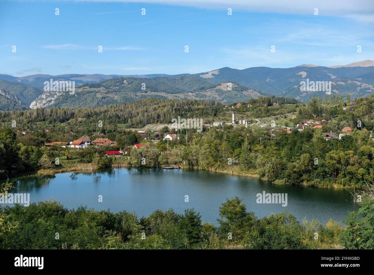 Vue panoramique sur un lac serein près de Petrosani, Roumanie, entouré de verdure luxuriante et de montagnes lointaines sous un ciel bleu clair. Banque D'Images