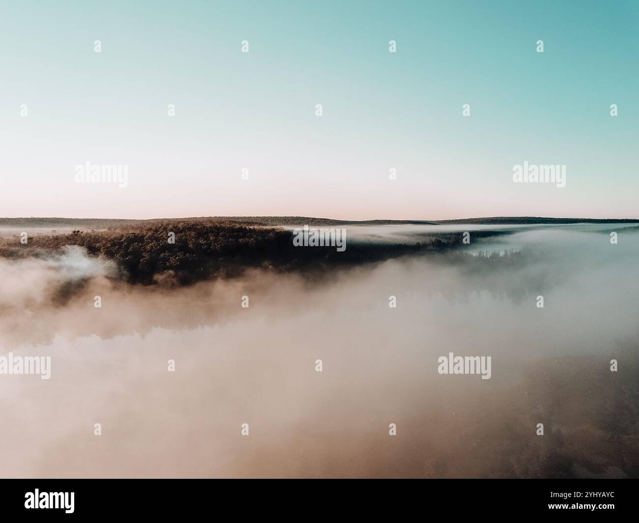 La couverture nuageuse basse commence à se dégager sur le lac et la forêt de pins à l'aube alors que le soleil commence à se lever. Vue du dessus des nuages. Banque D'Images