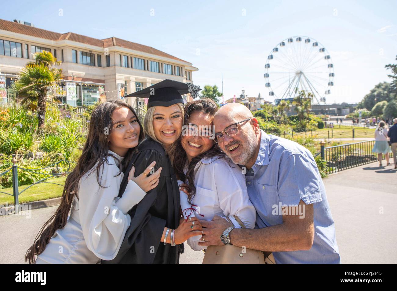 Un joyeux groupe familial de quatre personnes célèbre la remise des diplômes en plein air avec une grande roue en arrière-plan, Bournemouth, Dorset, Royaume-Uni Banque D'Images