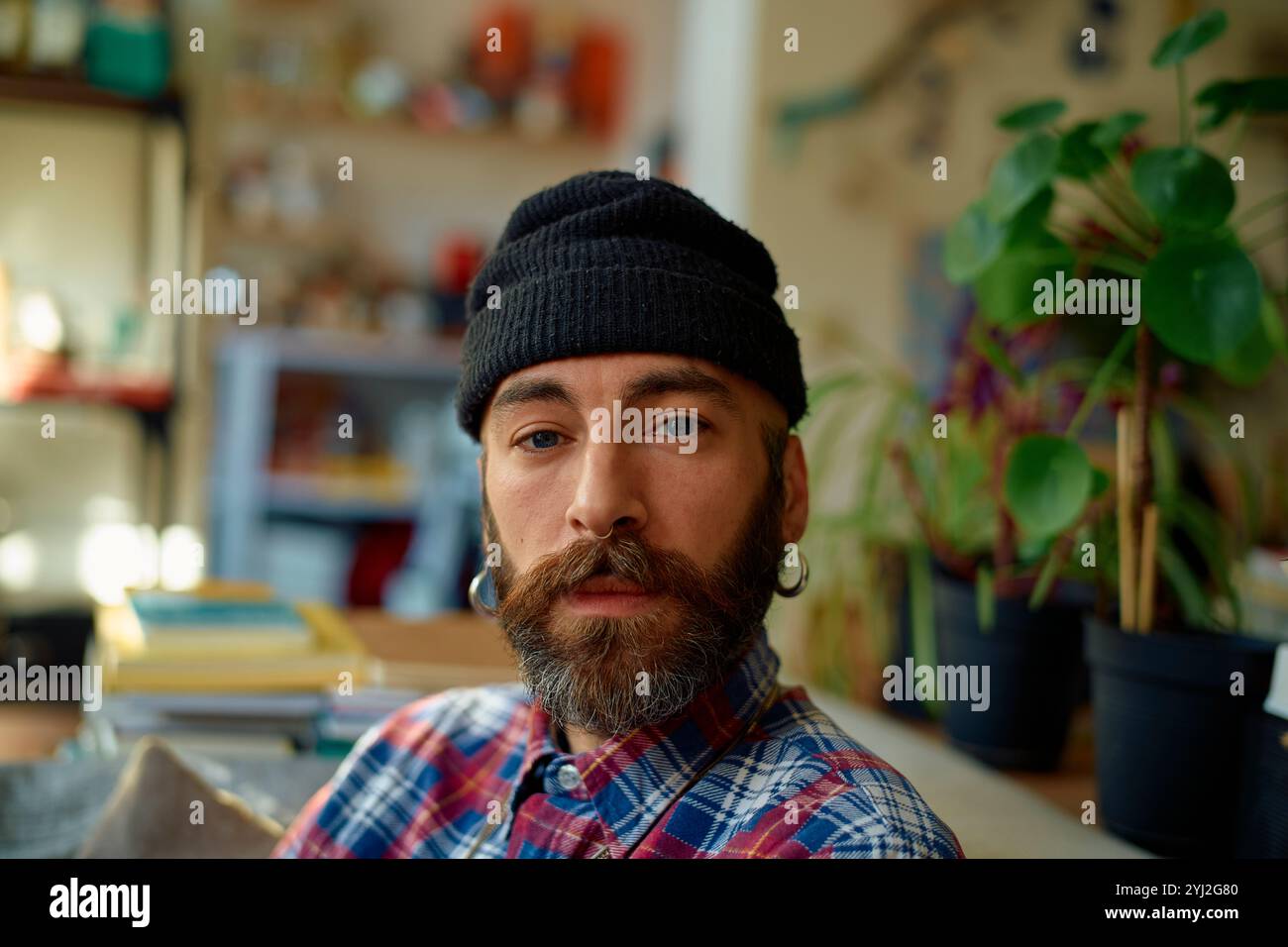 Homme barbu élégant dans un bonnet beanie pose décontractée dans une pièce vibrante décorée de plantes d'intérieur. Banque D'Images