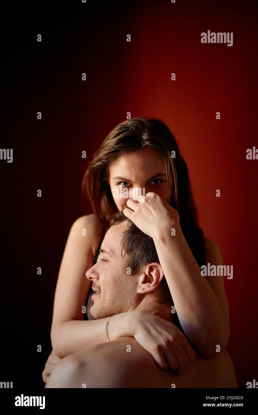 Femme souriant avec une main sur son visage tout en embrassant un homme par derrière sur un fond rouge. Banque D'Images