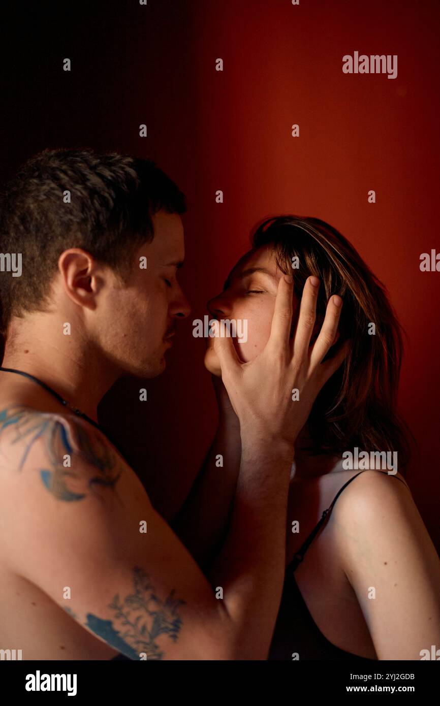 Un homme tatoué touche doucement le visage d’une femme alors qu’elle partage un moment intime sur fond rouge. Banque D'Images