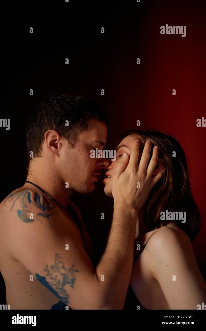 Moment intime d'un homme tatoué tenant tendrement le visage d'une femme alors qu'ils sont sur le point de s'embrasser, avec un fond rouge foncé. Banque D'Images