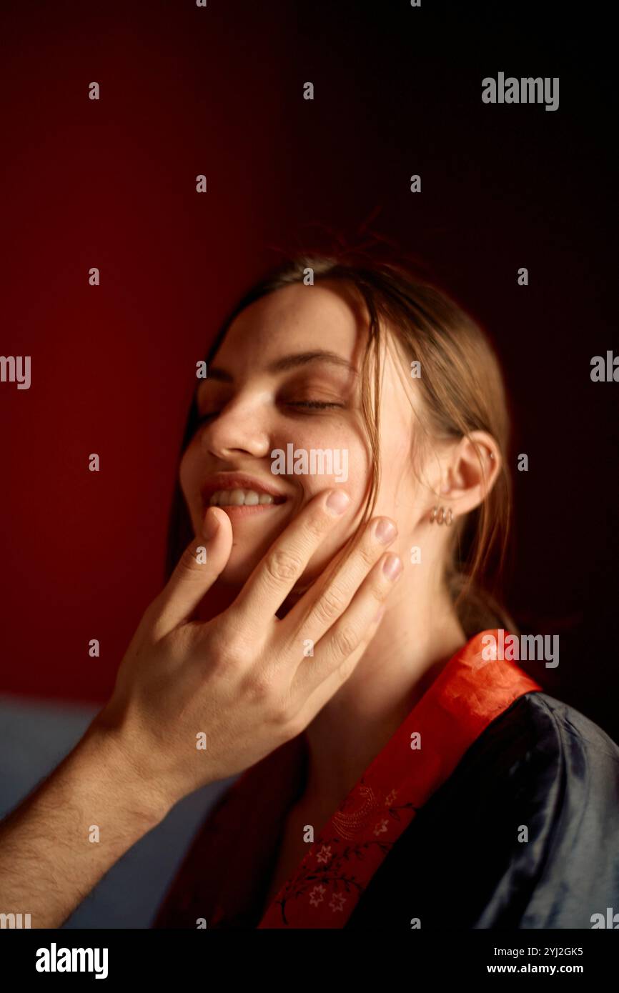 Femme souriante avec les yeux fermés appréciant un toucher doux à son visage dans une pièce légèrement éclairée. Banque D'Images