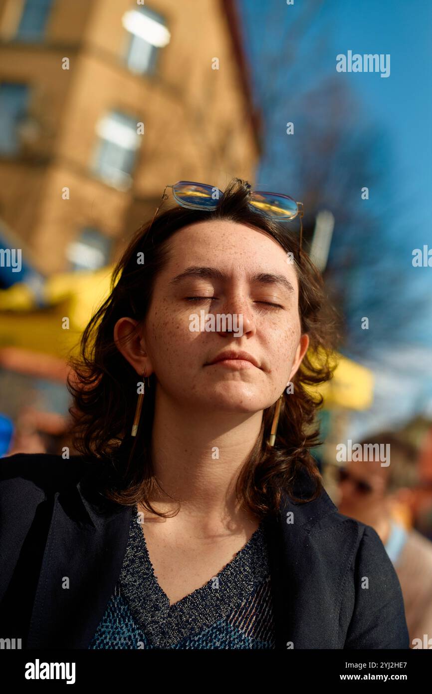Une jeune femme avec les yeux fermés et des lunettes de soleil sur la tête se tient dehors, se prélassant dans la lumière du soleil. Banque D'Images